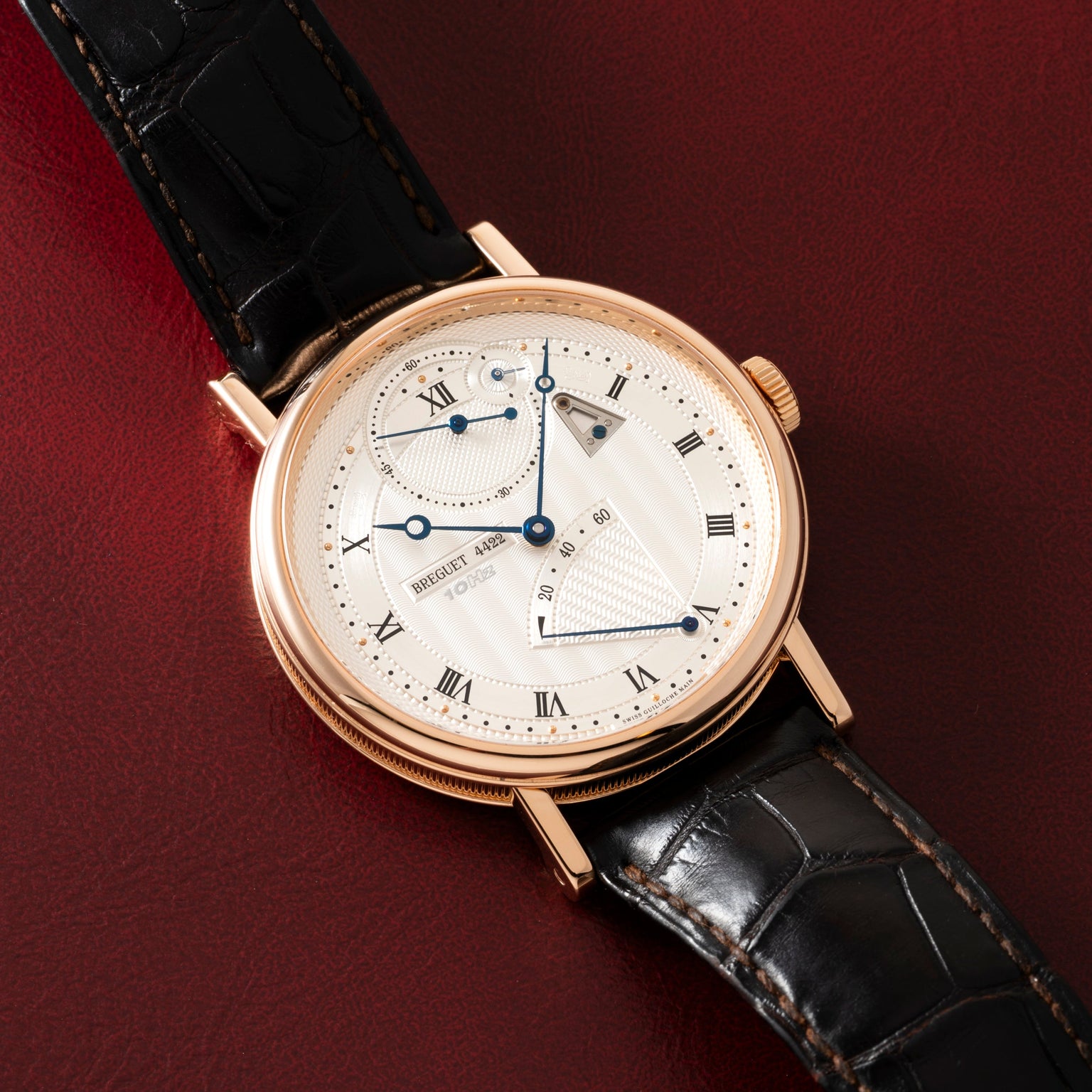 Rose gold watch with cream dial, Roman numerals, chronograph subdials, and black leather strap on burgundy background.