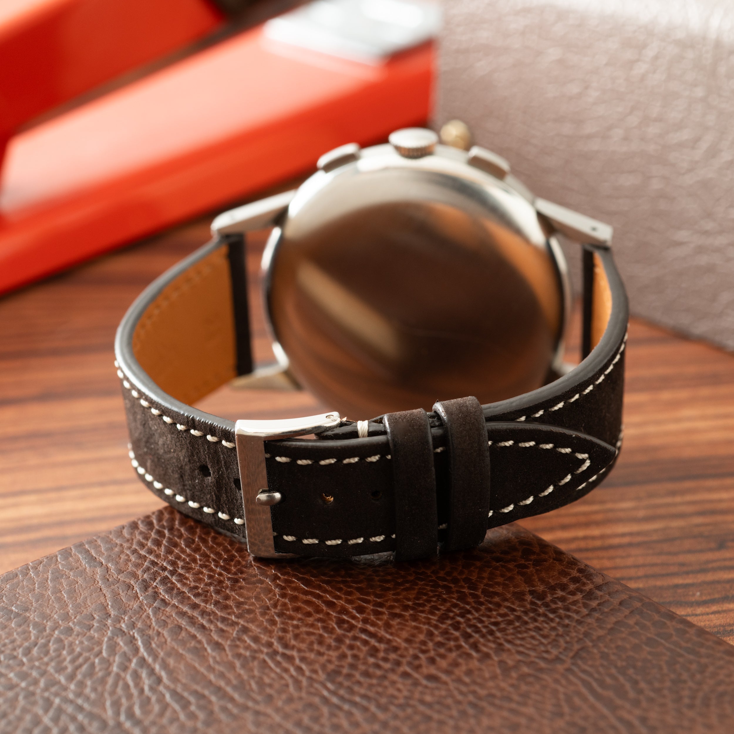 Stainless steel chronograph watch with dark brown leather strap and white stitching, displayed on wooden surface.