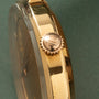 Gold-toned watch crown with engraved emblem, viewed from the side against a green background.