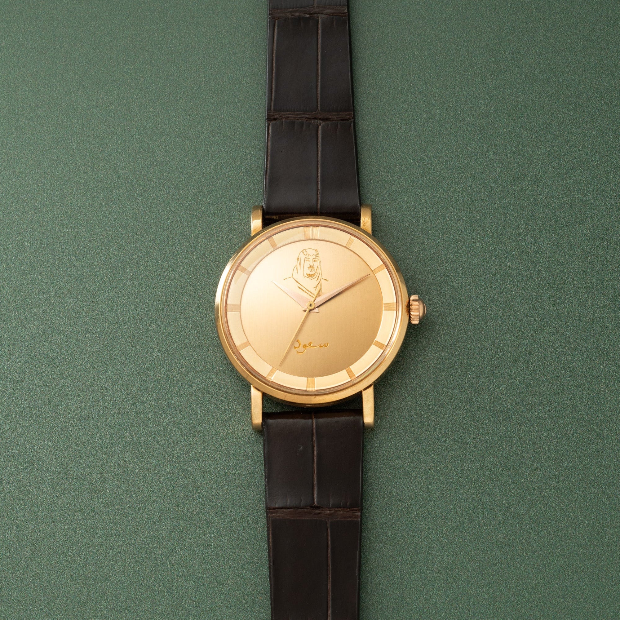 Gold-toned wristwatch with cream dial, portrait engraving, and black leather strap on green background.