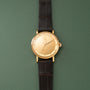 Gold-toned wristwatch with cream dial, portrait engraving, and black leather strap on green background.