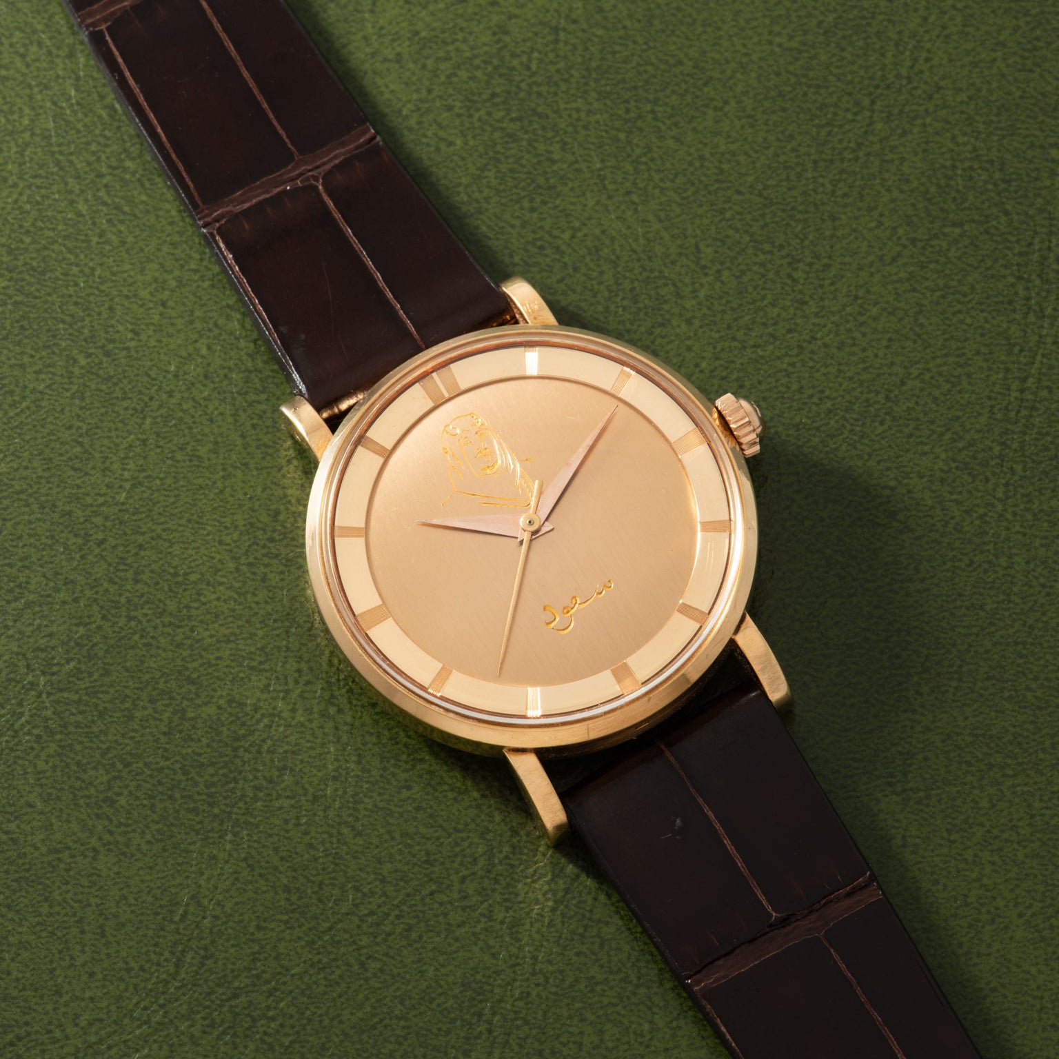 Gold-tone watch with champagne dial, Arabic numerals, and dark brown crocodile leather strap on green background.