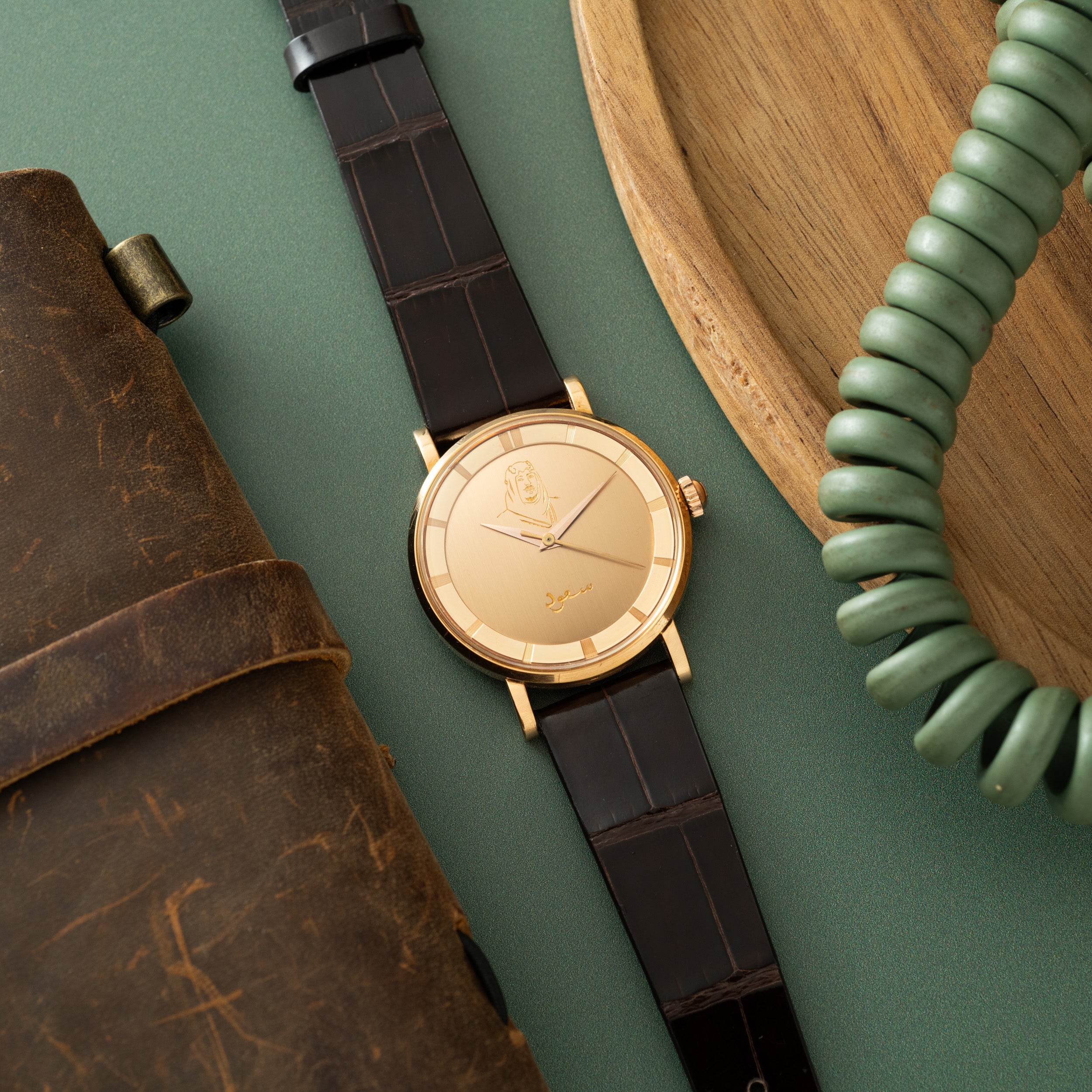 Gold-toned round watch with dark leather strap, displayed with brown leather pouch and green beaded bracelet on teal backgrou
