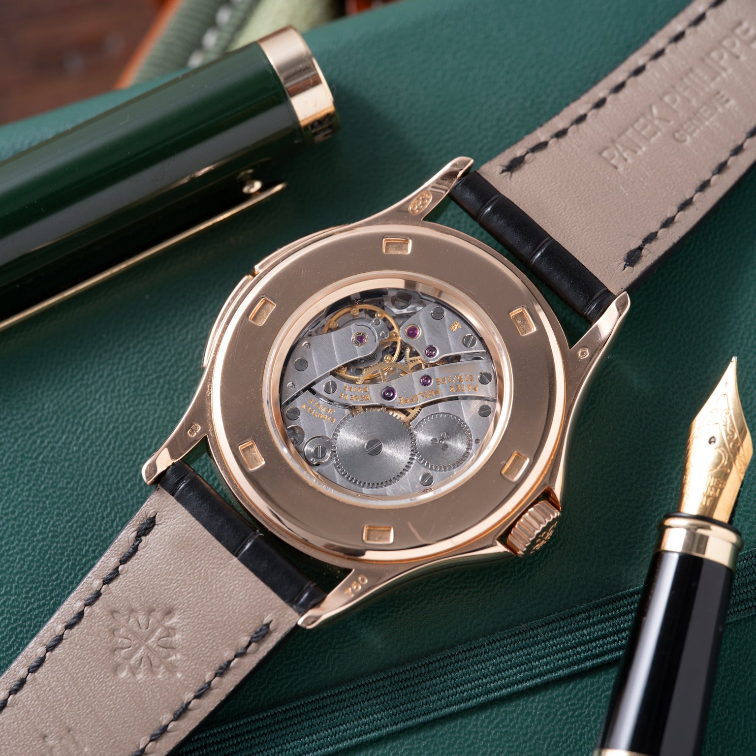Rose gold watch with skeletonized dial showing mechanical movement, black leather strap, displayed on green fabric with pen.