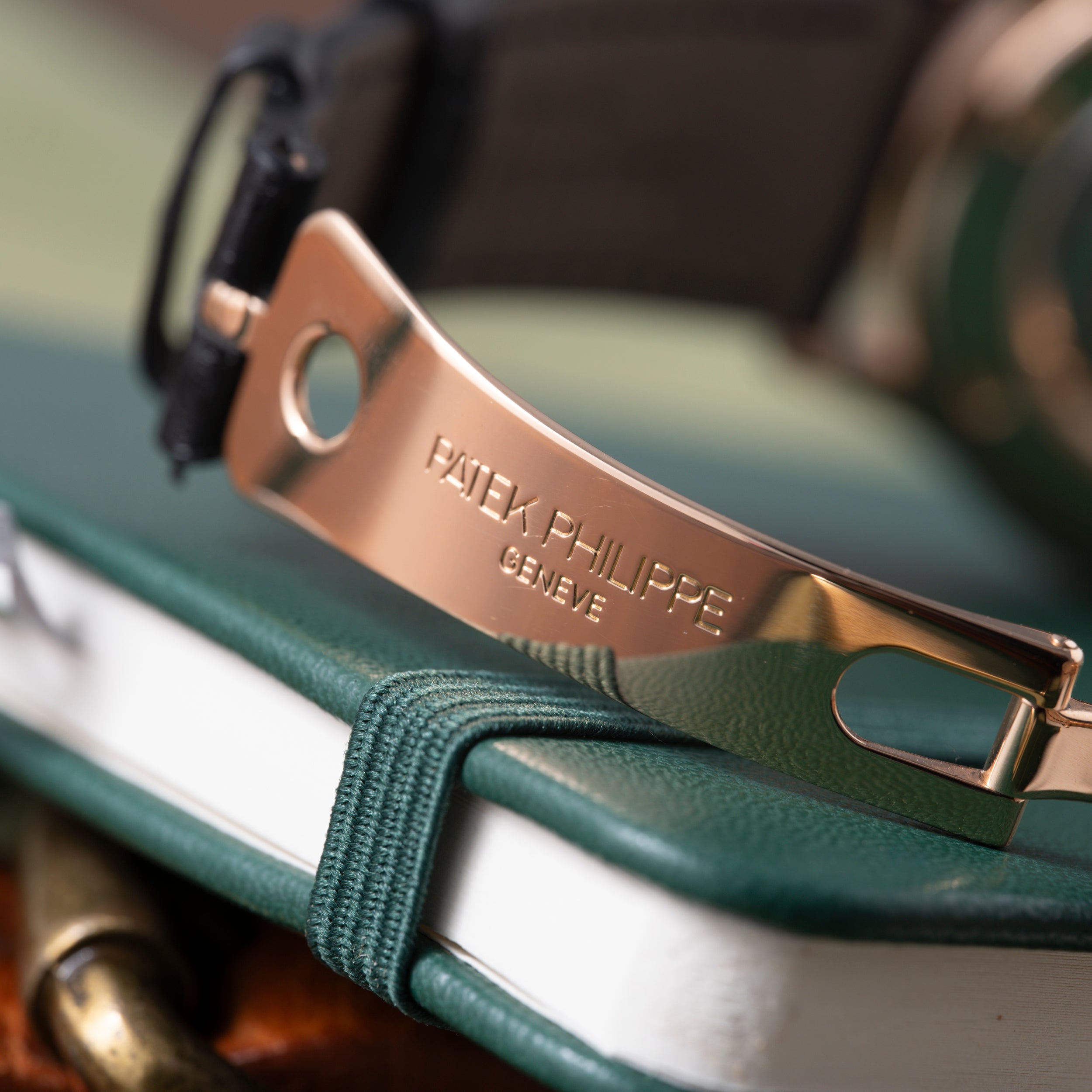 Rose gold watch clasp engraved with "PATEK PHILIPPE GENEVE" on green and white leather strap.