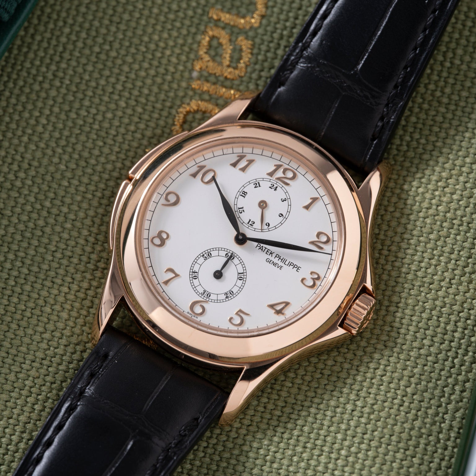 Rose gold watch with white dial, Arabic numerals, two subdials, and black leather strap on green fabric background.