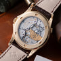 Gold-tone watch with exposed movement visible through caseback, dark leather strap with contrast stitching.