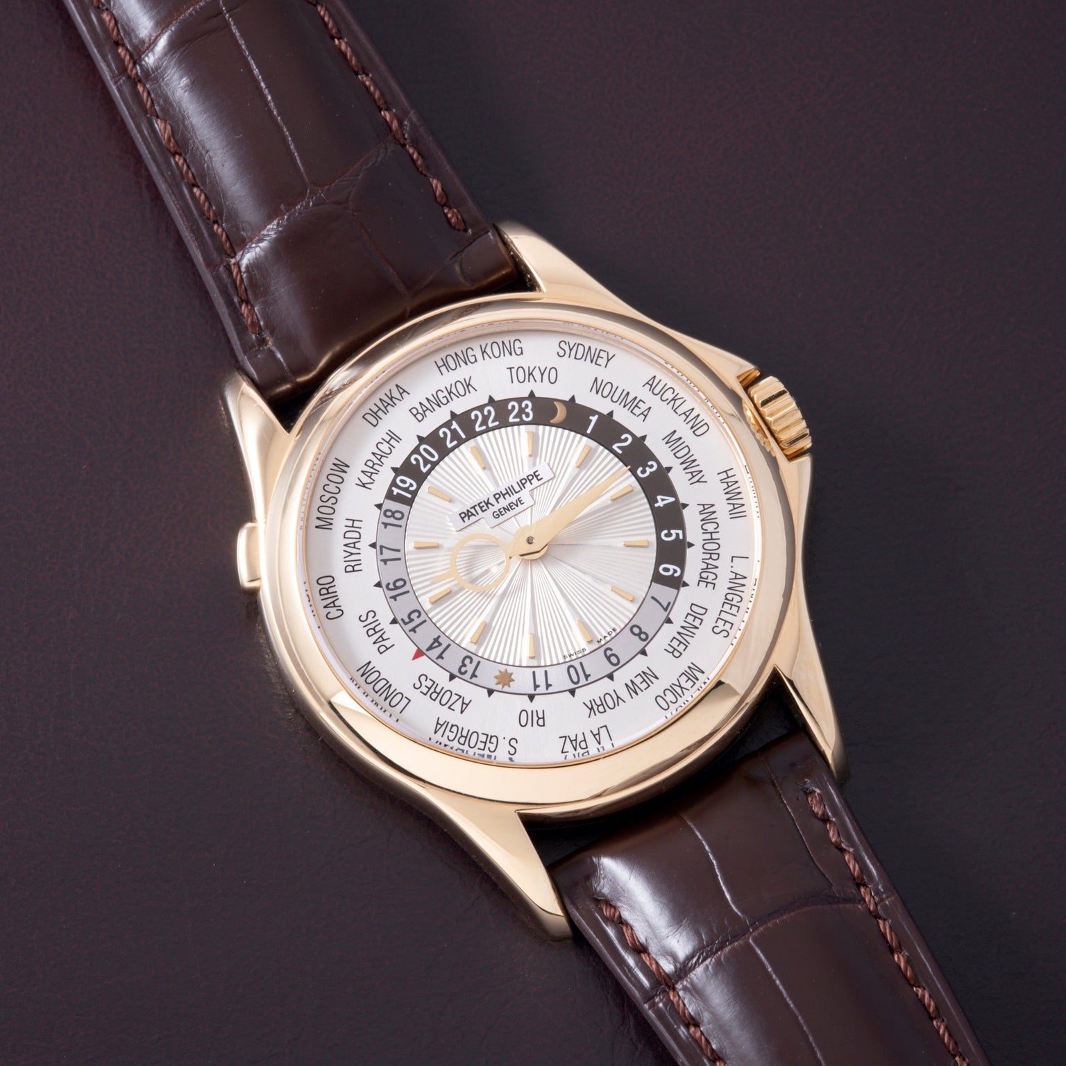 Gold watch with white dial, world city names around outer ring, dark leather crocodile-textured strap with orange stitching.