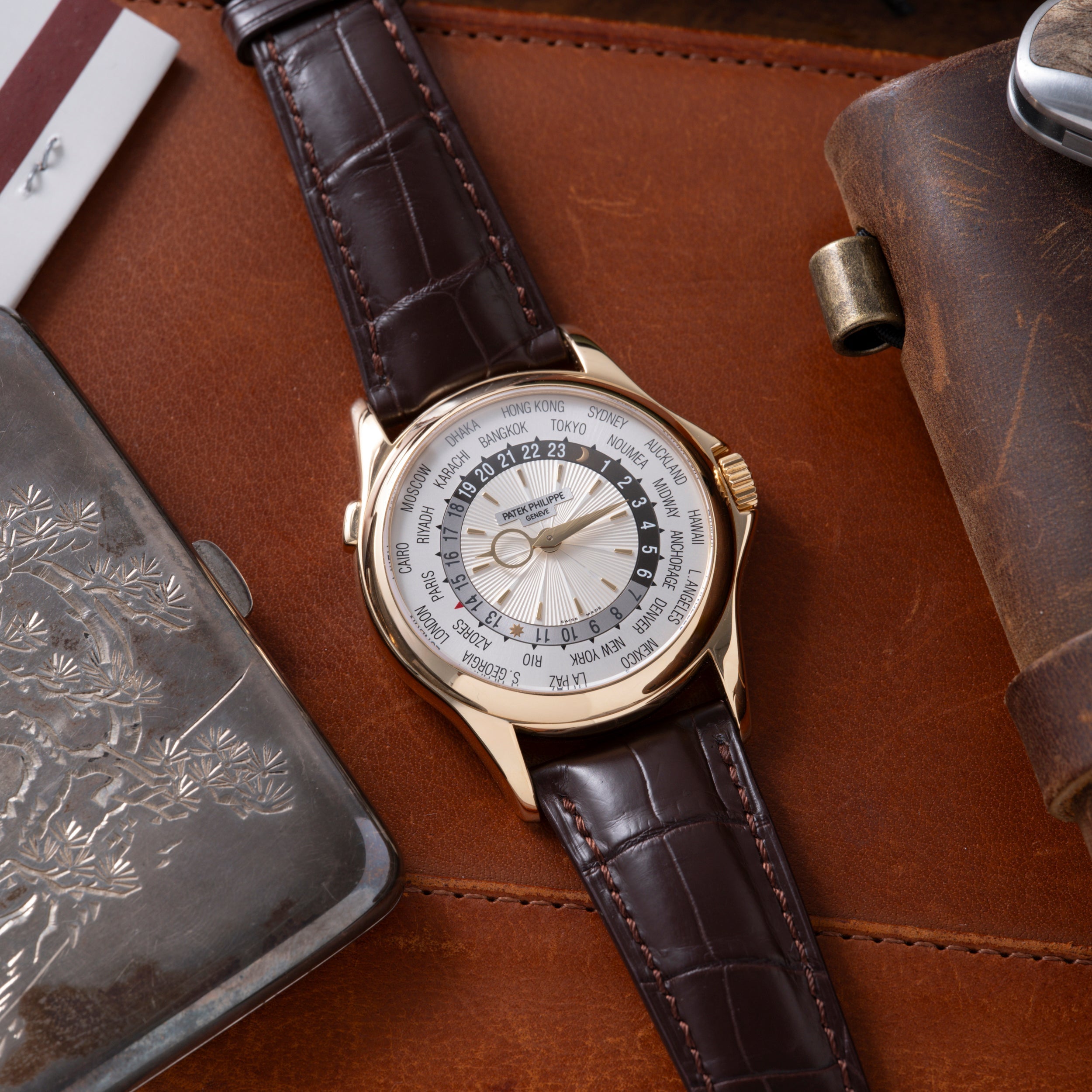 Gold-tone watch with white dial displaying world city names and 24-hour time zone ring, dark brown leather strap.