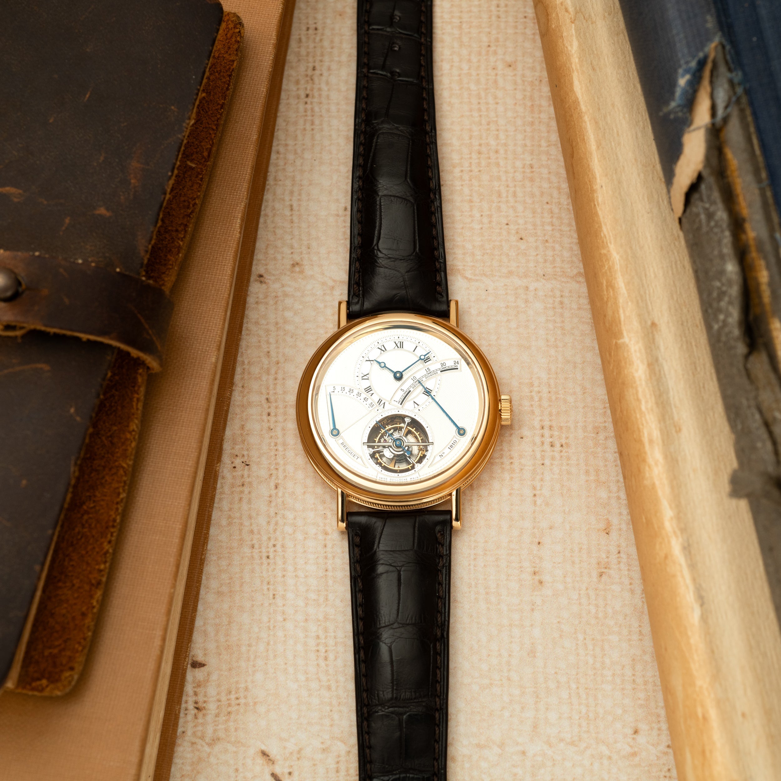 Gold-cased watch with white dial, Roman numerals, black crocodile leather strap, and visible tourbillon at bottom.