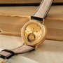 Gold-tone watch with engraved dial, visible tourbillon mechanism, and beige leather strap on wooden surface.