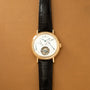 Gold-tone watch with white dial, Roman numerals, black crocodile leather strap, and visible tourbillon mechanism at bottom.