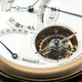 Luxury watch with cream dial, Roman numerals, blue hands, visible tourbillon mechanism in circular aperture, and gold-tone ca