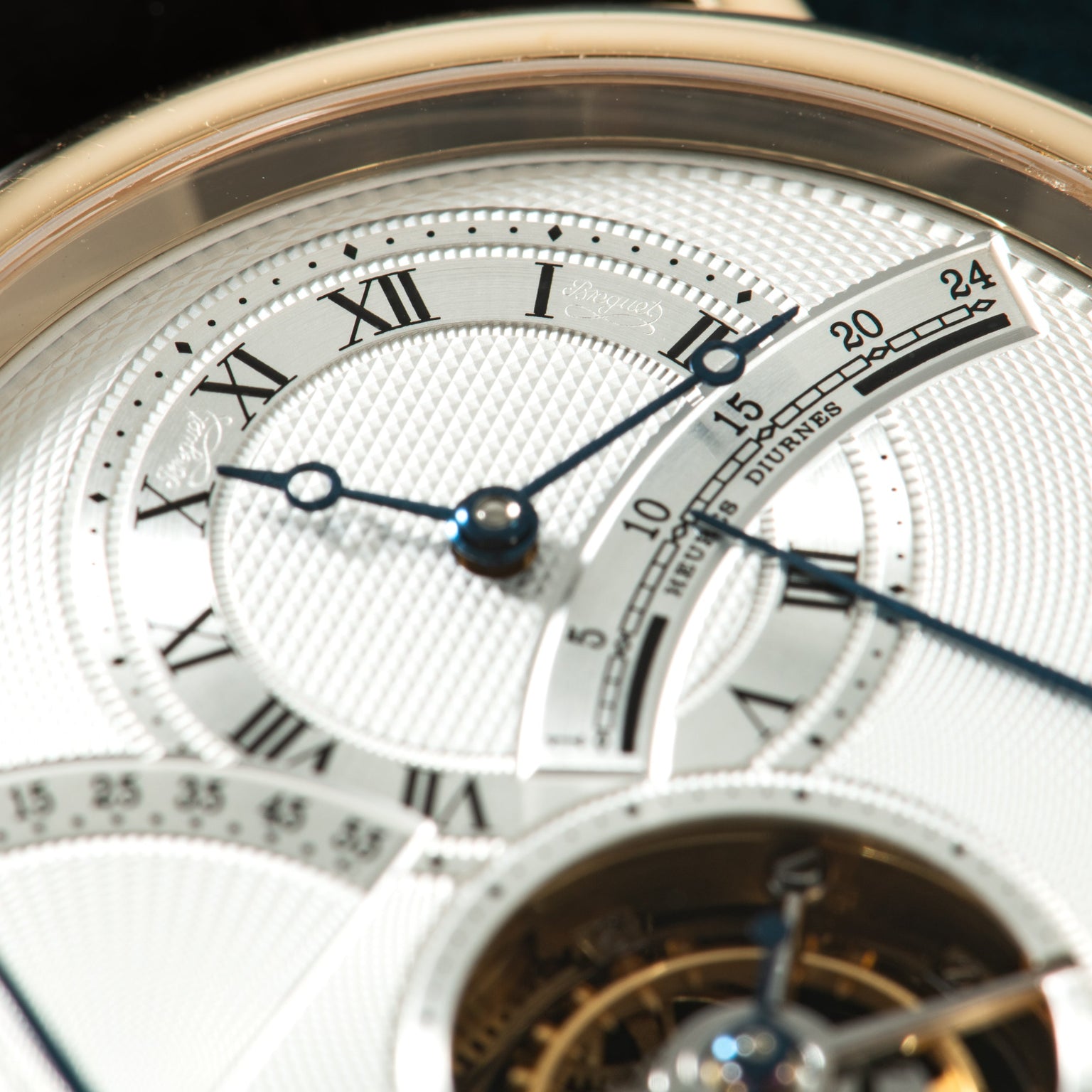 Gold-tone watch face with white dial, Roman numerals, blued hands, date window, and tourbillon visible at bottom.