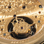 Gold watch movement with ornate engraved plate, visible tourbillon cage, jeweled bearings, and mechanical gears through displ