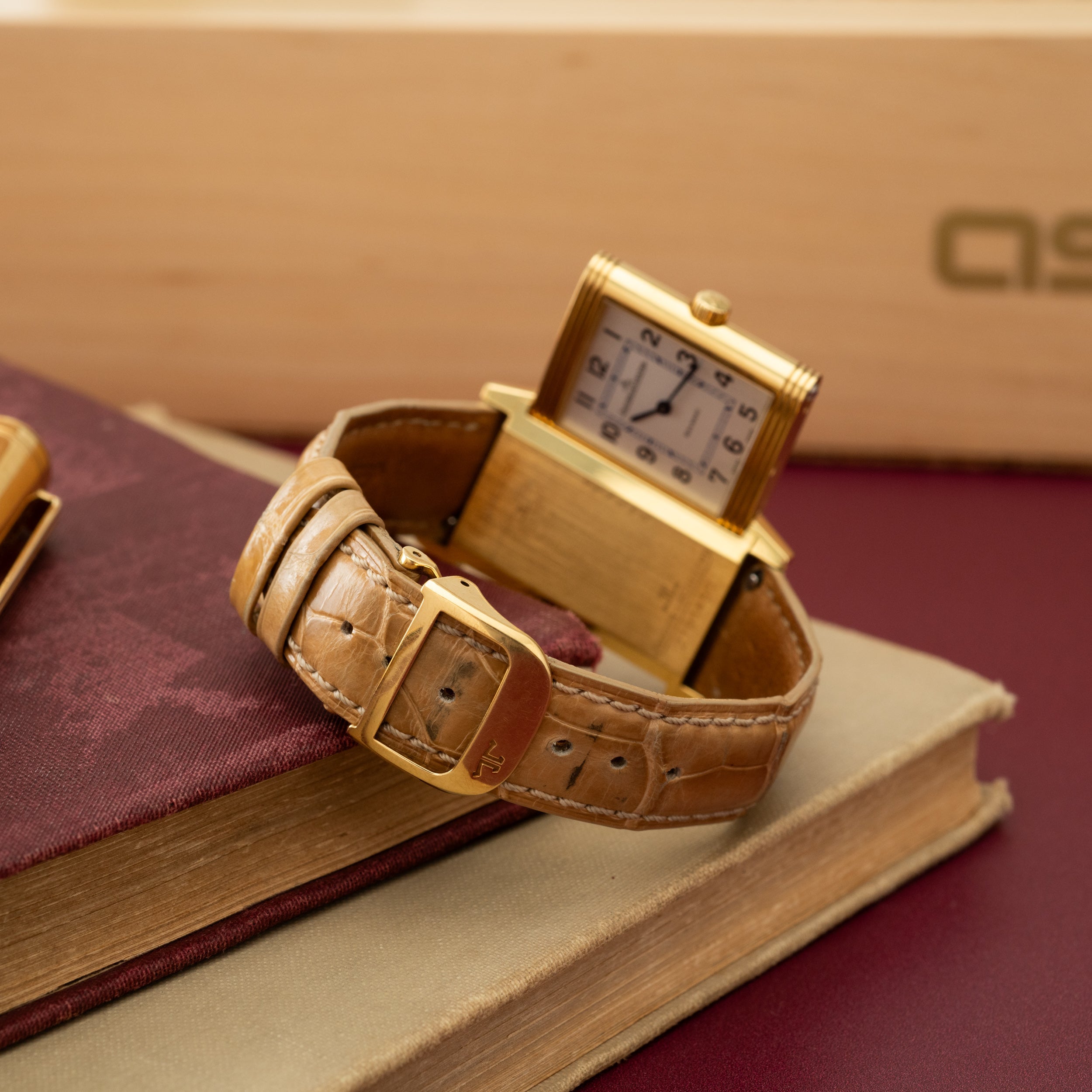 Gold rectangular watch with cream dial and Arabic numerals on tan crocodile leather strap, displayed on burgundy fabric.