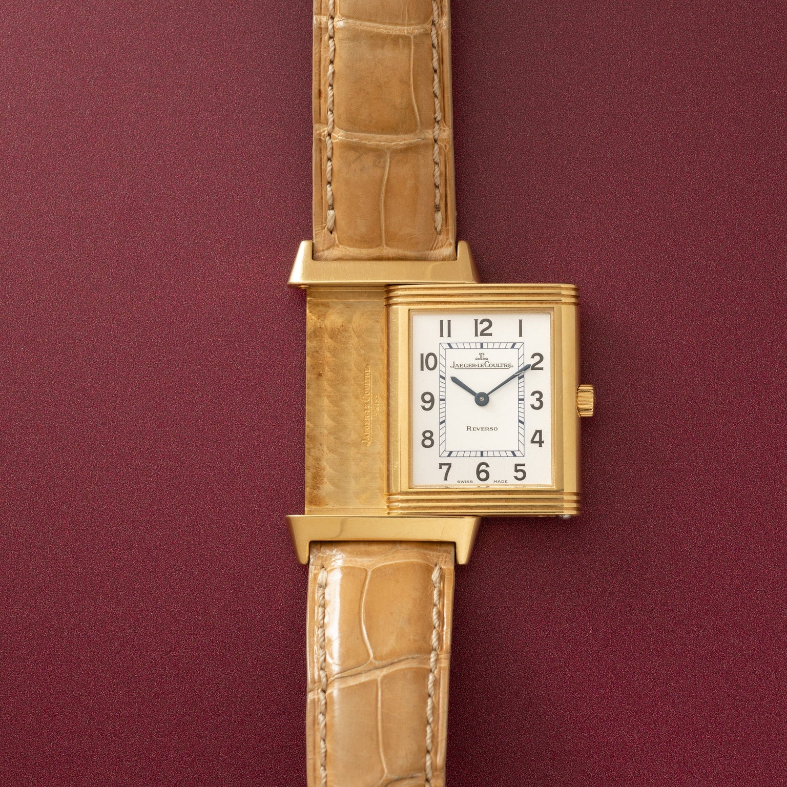 Gold-toned rectangular watch with cream dial, Arabic numerals, and tan crocodile leather strap against burgundy background.
