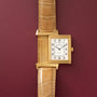 Gold-toned rectangular watch with cream dial, Arabic numerals, and tan crocodile leather strap against burgundy background.