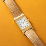 Gold-tone rectangular watch with white dial, Arabic numerals, and tan crocodile leather strap on yellow background.