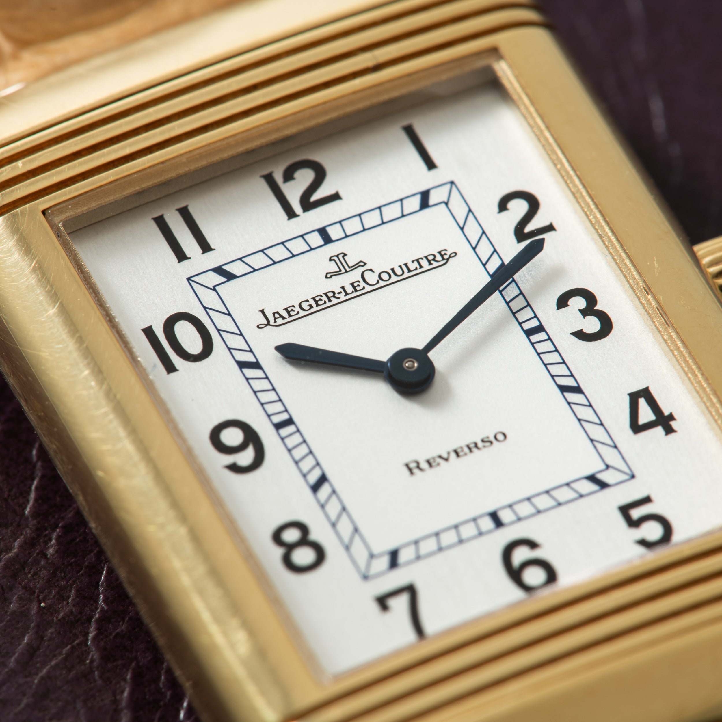 Gold-toned rectangular watch with white dial, black Arabic numerals, and black hands displaying Jaeger-LeCoultre branding.