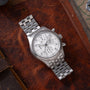 Stainless steel chronograph watch with white dial, multiple subdials, and linked metal bracelet on brown leather surface.
