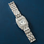 Silver-tone square watch with diamond-set bezel, Roman numeral dial, and multi-link metal bracelet on blue background.