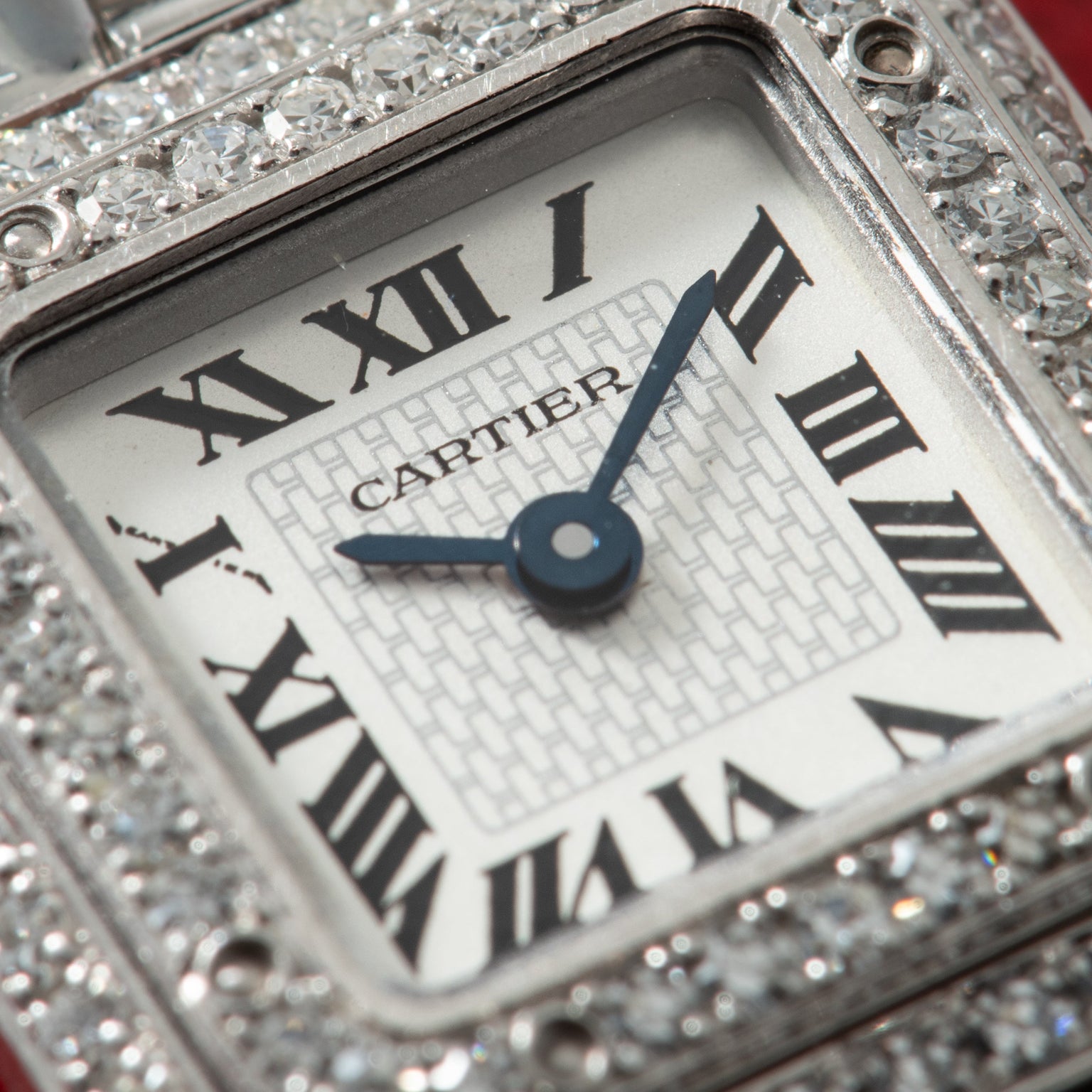 Cartier watch face with Roman numerals and blue hands, surrounded by diamond-set bezel on metal case.