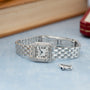 Silver-toned square watch with diamond-set bezel, Roman numeral dial, and linked metal bracelet on light blue surface.