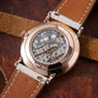 Rose gold watch with skeletonized dial showing visible gears and jewels, paired with cognac and cream leather strap.