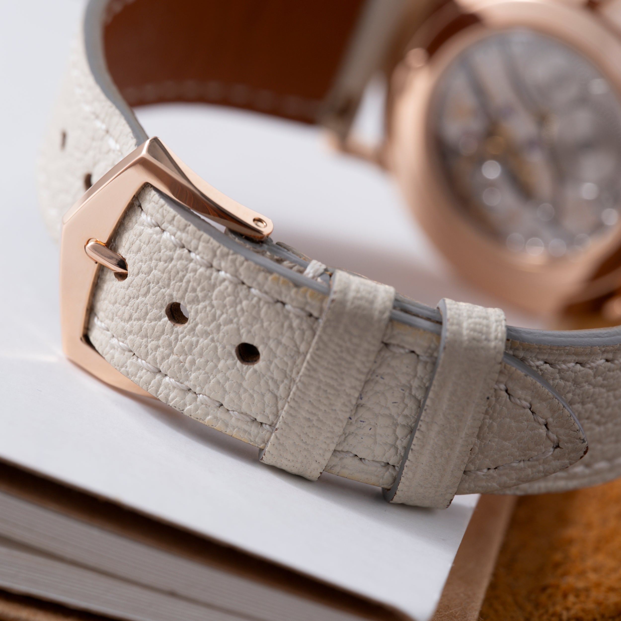 Rose gold watch with cream leather strap and rectangular buckle, shown at an angle with blurred watch face visible in backgro