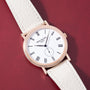 Rose gold watch with white dial, Roman numerals, subdial, and white leather strap on burgundy background.