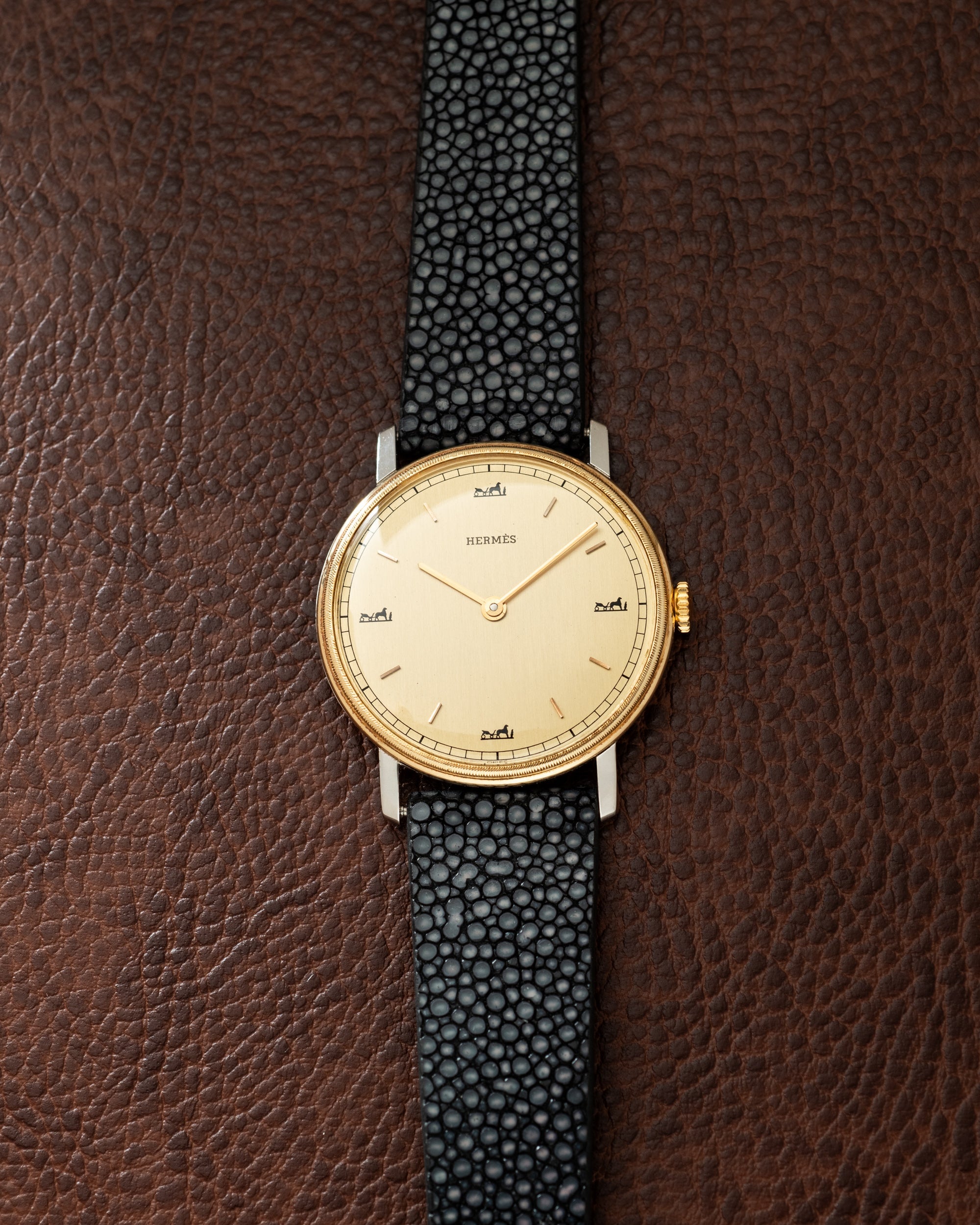 Hermès watch with gold-tone round case, cream dial, and black textured leather strap on brown leather background.