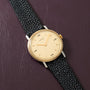 Hermès watch with cream dial, gold-tone case, Arabic numerals, and black textured leather strap on purple background.
