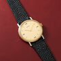 Hermès watch with cream dial, gold-tone case, and black textured leather strap on red background.