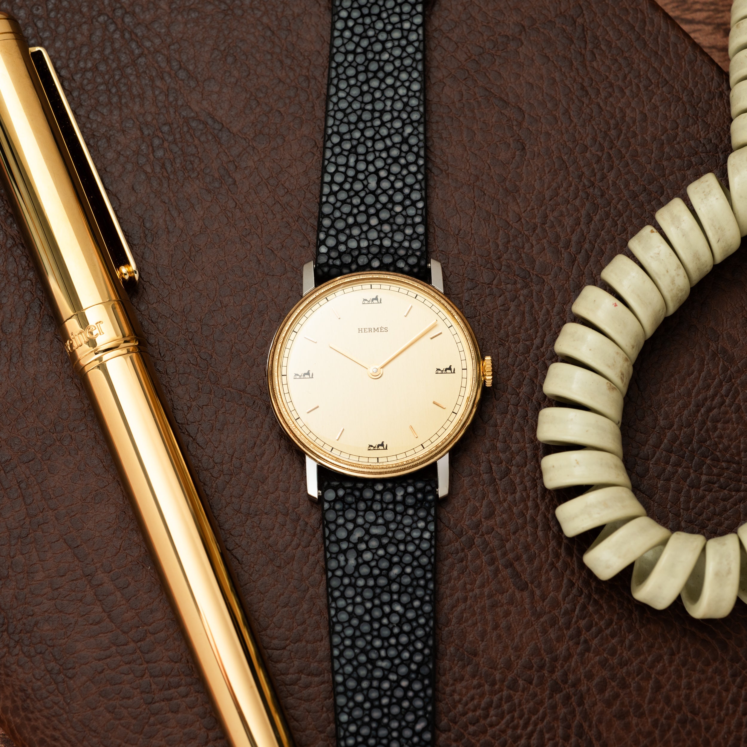 Gold-tone round watch with cream dial, black leather strap, flanked by gold pen and cream beaded bracelet on brown leather ba