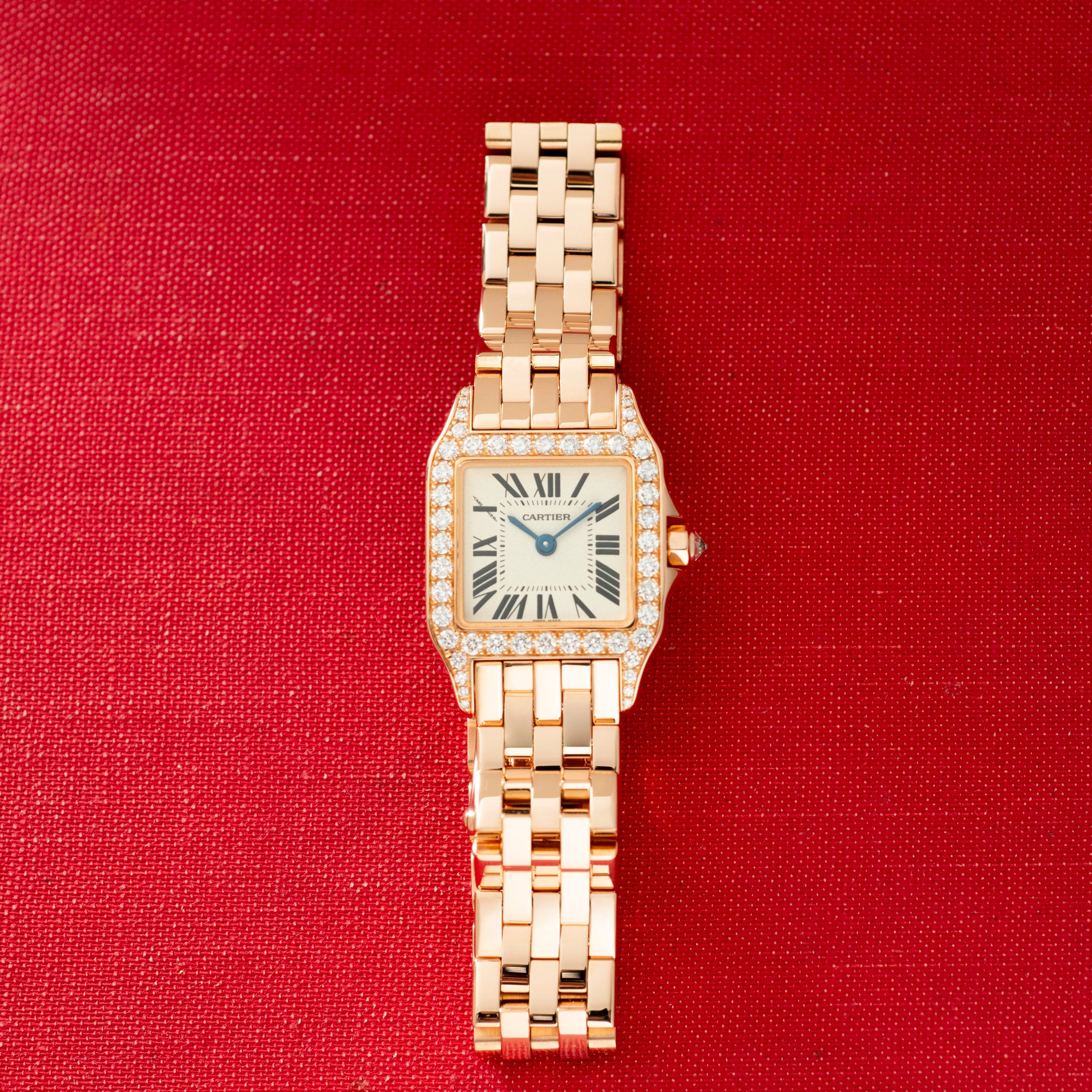 Rose gold watch with square face, diamond-set bezel, Roman numeral dial, and linked bracelet on red background.