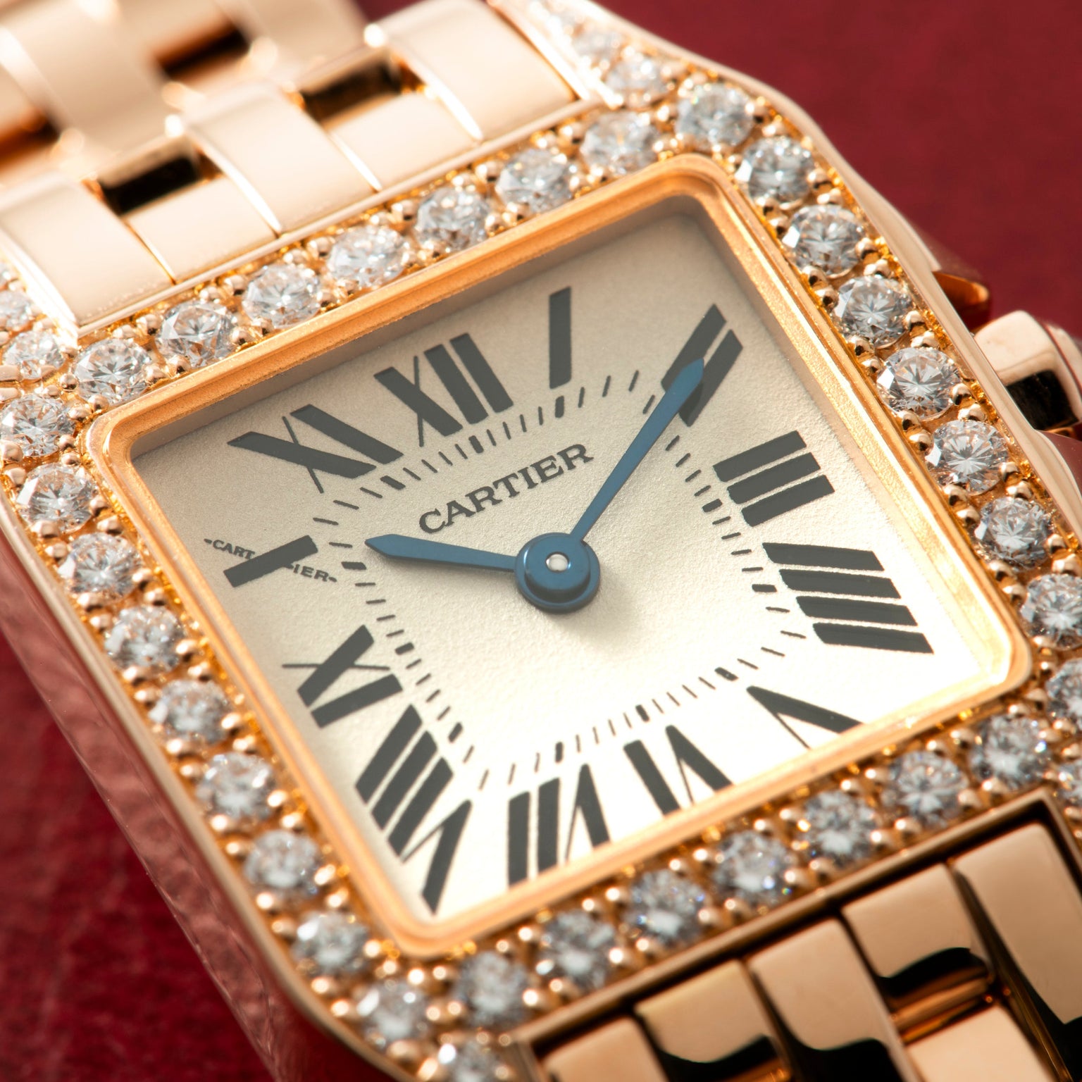 Rose gold square watch with diamond bezel, cream dial, Roman numerals, blue hands, and matching bracelet.