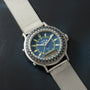 Silver-toned watch with blue dial, yellow hour markers, dual analog and digital displays, and white mesh band on dark surface