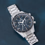 Stainless steel chronograph watch with black dial, tachymetre bezel, and metal bracelet on blue background.