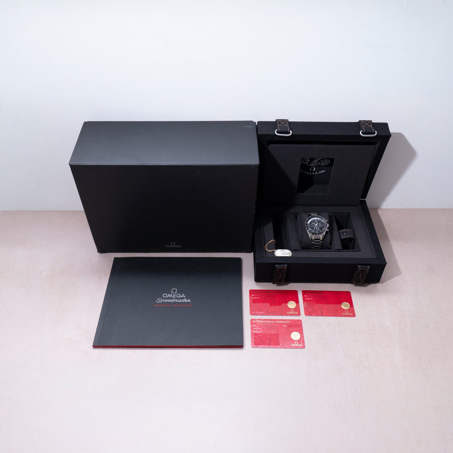 Omega Speedmaster watch with chronograph dial in black case, displayed with black presentation box, gray documentation folder