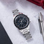 Omega Speedmaster chronograph watch with black dial, tachymetre bezel, steel bracelet, and orange hour markers on marble surf