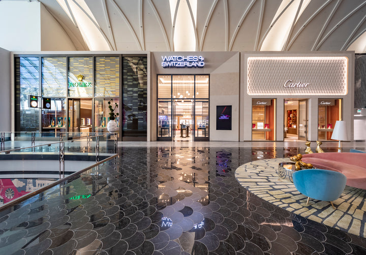 Mall with Rolex, Watches of Switzerland, and Cartier storefronts, glossy floor, and colorful seating in foreground.