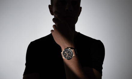 A silhouetted man wearing a black shirt shows off a wristwatch in dramatic lighting.