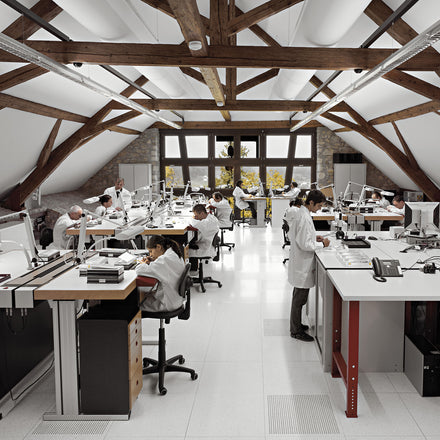 A workroom with white benches and craftspeople in white coats looking at watches and gears