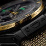 Black ceramic watch with gold bezel engraved with text, green dial accents, and matching gold-tone mesh bracelet.