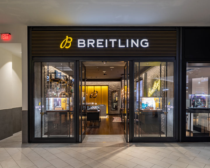 Breitling store with illuminated sign, glass entrance, and watch displays inside a shopping mall.