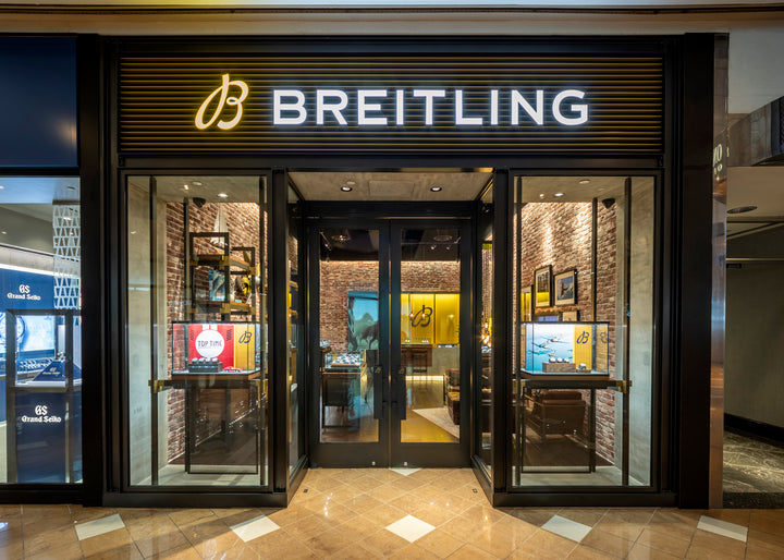 Breitling store entrance with illuminated logo, glass doors, and watch displays inside.