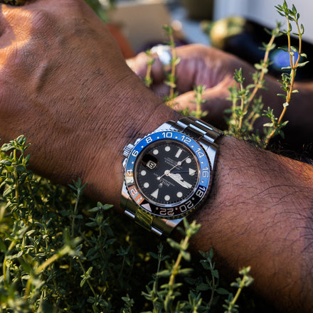 Close-up of a wrist wearing a watch with a blue bezel, surrounded by green plants.