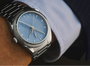 Silver watch with blue face on a wrist, wearing a light blue shirt.