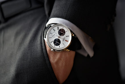 A person's hand in a pocket of a suit, the wrist wears a Czapek watch with a white dial and two black subdials